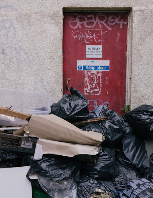 Side view of a small, light blue utility truck parked on a paved street, loaded with various types of waste materials including cardboard boxes, black plastic bags, and other debris. The truck's cargo