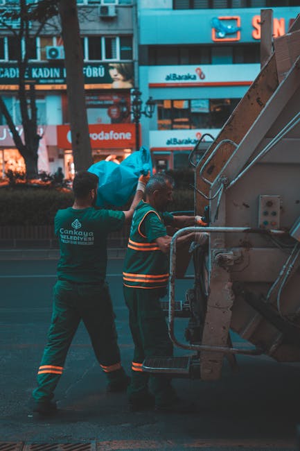 Two waste collection workers are engaged in rubbish removal on an urban street, standing beside a large industrial waste container. One worker, wearing a dark green uniform with reflective orange stri