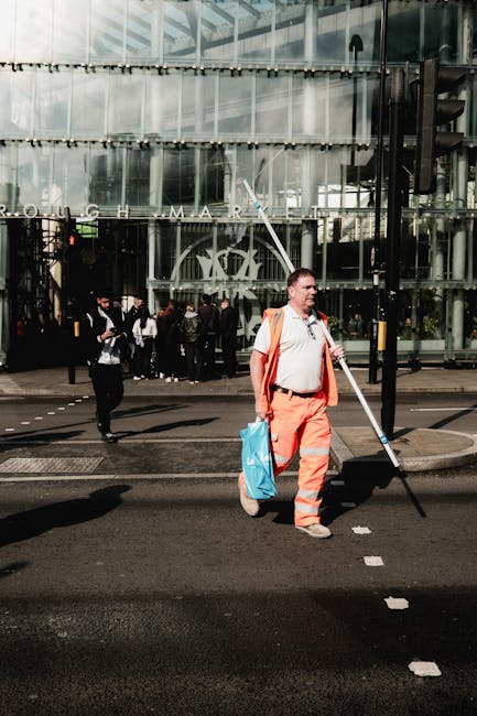 A street scene outside a modern glass office building with a large, partially reflective facade. In the foreground, a man dressed in a white shirt and bright orange safety trousers, holding a long pol