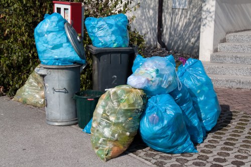 A collection of black, grey, and green rubbish bins filled with bags of household waste sits on a paved driveway adjacent to a residential building. Several large blue plastic bags and clear bags containing mixed waste are placed on the ground, some leaning against the bins and others stacked nearby. The background includes a section of flowering shrubbery and a concrete staircase leading to the house entrance, with a fence and a small garden area partially visible. The scene depicts a typical rubbish collection setup for a house clearance service in Pinner, with waste awaiting removal. The textures of the plastic bags and the smooth surfaces of the bins contrast with the paved and garden surroundings, illustrating a scene commonly managed by companies like House Clearance Pinner as part of local rubbish clearance tasks.
